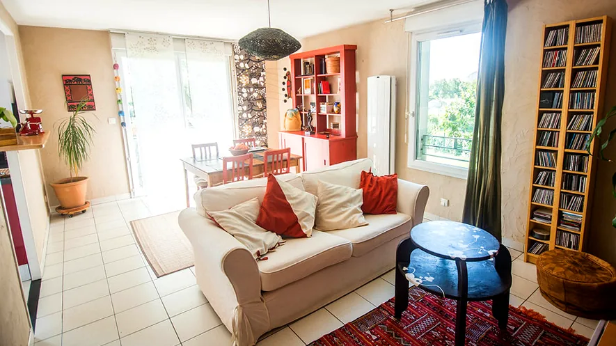 Vrai séjour d'appartement : canapé beige, table basse, tapis rouge et coin repas près de la baie vitrée